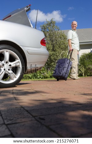 Senior man pulling suitcase on wheels from parked car boot on driveway, smiling, side view, portrait (surface level)