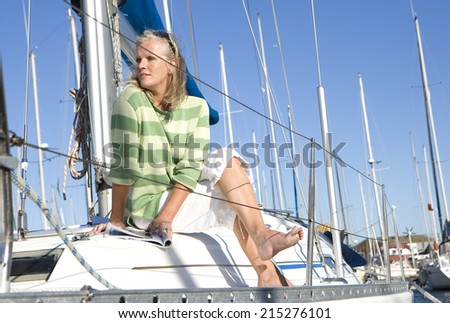 Mature woman, in green striped jumper, sitting on deck of yacht moored at harbour jetty, legs crossed at knee, looking away, thinking
