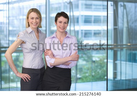 Two businesswomen standing beside large window in office, arm in arm, smiling, portrait
