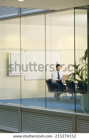 Solitary businessman working on laptop at conference table in boardroom, view through large office window
