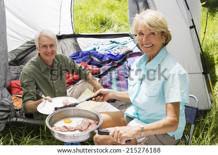 Senior woman serving husband fried breakfast on camping trip, man sitting inside tent, smiling, portrait