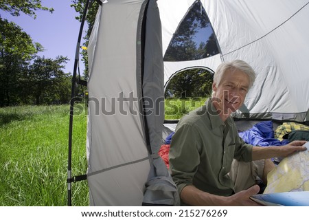 Senior man sitting inside tent in woodland clearing, looking at map, smiling, side view, portrait