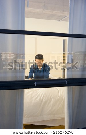Young man using laptop on bed, view through window from outside