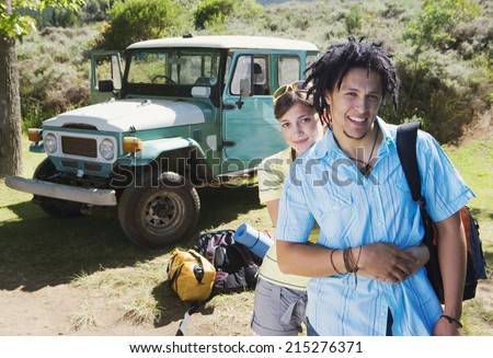 Young couple standing near parked jeep at start of camping holiday, woman embracing man, smiling, portrait