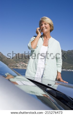 Senior woman standing beside parked convertible car on clifftop overlooking bay, using mobile phone, smiling