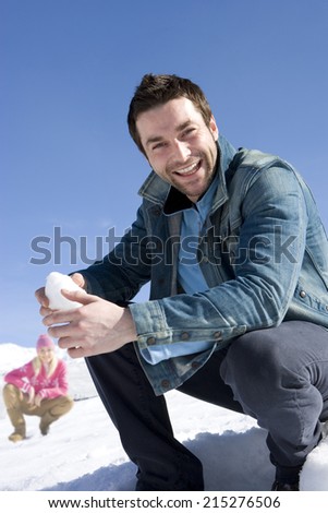 Young man crouching in snow field, holding snow ball, smiling, portrait, young woman in background