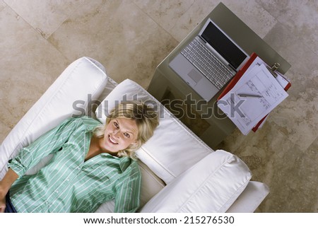 Woman lying on sofa beside laptop on coffee table at home, hands behind head, smiling, overhead view