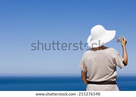 Senior woman taking photograph with digital camera of Atlantic Ocean from clifftop, rear view