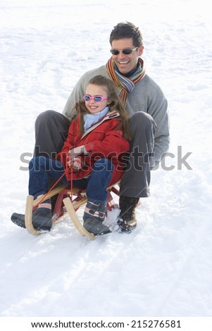Father and daughter riding sled down snow slope, smiling, low angle view