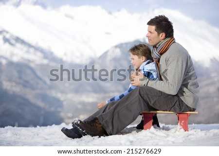 Father and son sitting on sled in snow field, side view, mountain range in background