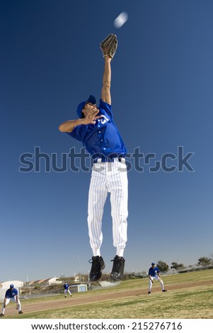 Baseball player, in blue uniform, jumping up to catch ball in protective glove during competitive game (surface level, tilt)
