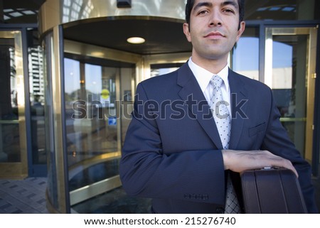 Businessman standing in front of revolving door, carrying briefcase, smiling, front view, portrait