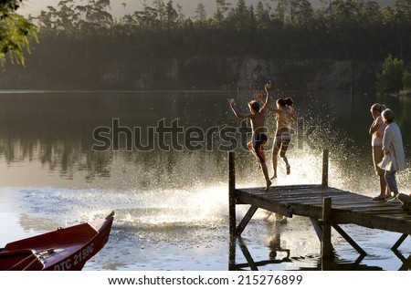 Multi-generational family standing on jetty at sunset, children (7-10) jumping into lake, side view
