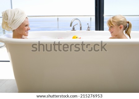 Mother and daughter (6-8) in bath outdoors, smiling at each other, side view