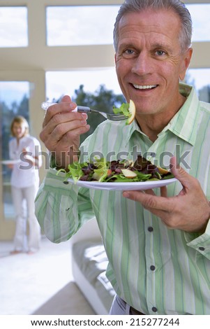 Senior man eating salad on plate at home, mature woman in background, focus on man, smiling, portrait
