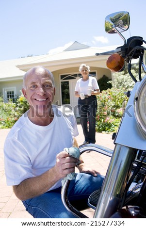 Senior man polishing motorbike on driveway, crouching down, senior woman serving drinks on background, smiling, portrait