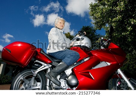 Senior woman sitting on red motorbike on driveway, holding crash helmet, side view, portrait, low angle view