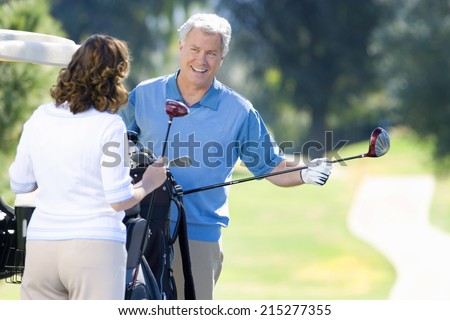Mature couple playing golf, man in blue tank top taking driver from golf bag, smiling