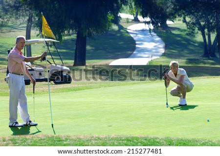Mature couple playing golf, woman lining up golf shot on putting green, man holding flag