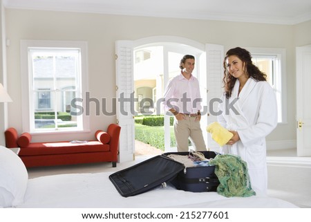 Young woman wearing white bath robe, packing suitcase on bed, man smiling in background