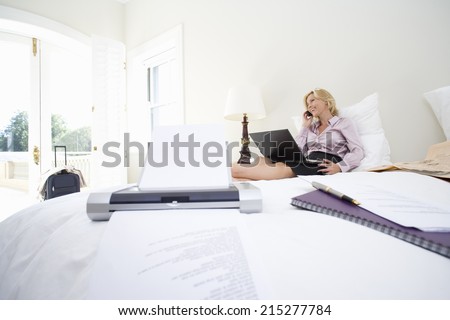 Mature businesswoman using laptop and mobile phone on bed, smiling, printer on bed in foreground