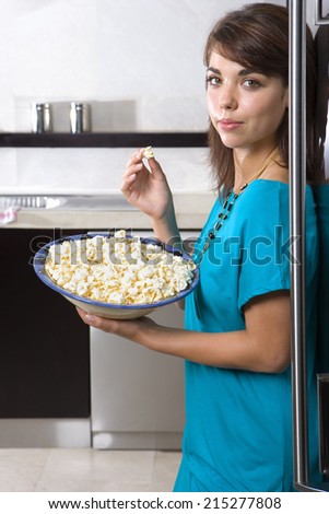 Young woman standing by fridge with bowl of popcorn, smiling, portrait