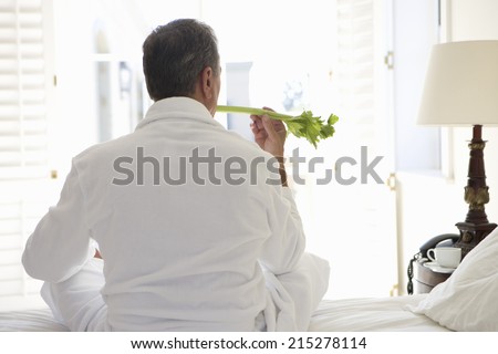 Senior man sitting on bed holding celery stick, rear view