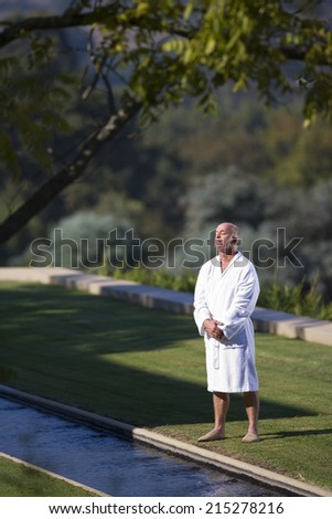 Mature man wearing white bath robe standing on lawn, eyes closed, elevated view