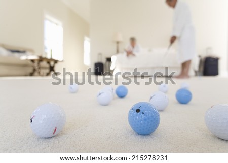 Senior couple in bedroom, man practising golf putt, focus on golf balls in foreground