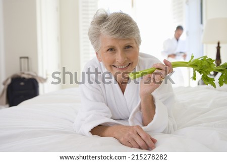 Senior woman lying on bed holding celery stick, smiling, portrait, senior man in background