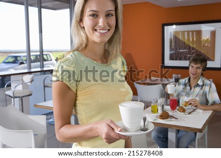 Young woman holding tea cup in cafeteria, smiling, portrait, young man smiling in background