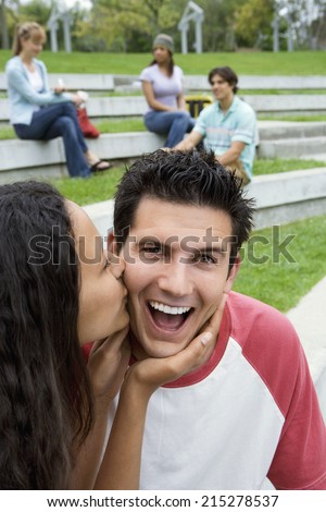 Teenage couple sitting near friends, girl kissing boy on cheek, smiling