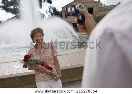Senior woman standing beside fountain, holding bouquet of red roses, smiling, man filming her with portable video recorder (tilt, differential focus)