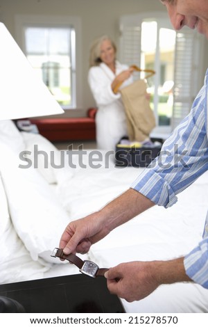 Mature man holding watch by bed, mature woman unpacking suitcase in background, smiling