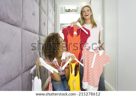 Two young woman holding up racks of clothes in corridor of change rooms in shop, smiling