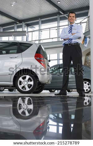 Car salesman standing near new cars in showroom, holding brochure, smiling, portrait (surface level)