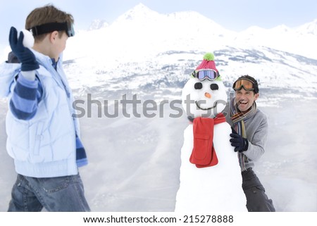 Boy throwing snowball at father standing by snowman in snow, smiling, mountain range in background