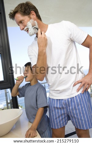Father shaving by son (6-8) brushing his teeth in bathroom, smiling, side view