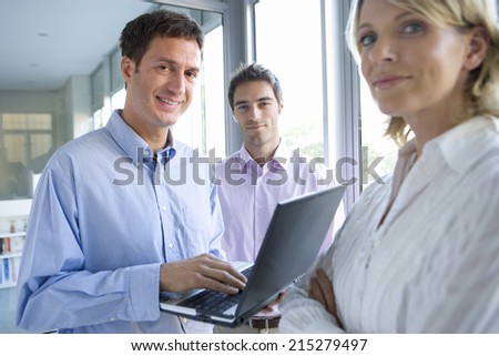 Businessman using laptop computer by colleagues standing by window, smiling, portrait