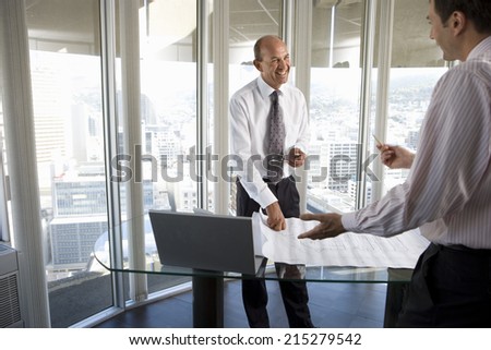 Mature businessman smiling at colleague in office, blueprint on desk