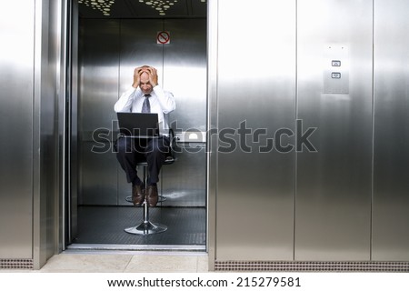 Mature businessman on stool in lift, laptop on lap, head in hands