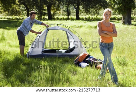 Young couple assembling tent on camping trip in woodland clearing, woman smiling, arms folded, portrait