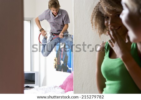 Two teenage girls (15-17) laughing at friend play 'air guitar' with tennis raquet in bedroom