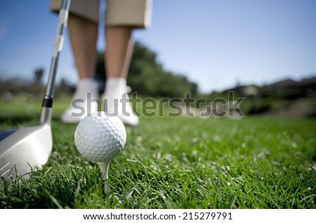 Mature woman preparing to tee off with driver on golf course, close-up, low section, focus on golf ball in foreground (surface level)