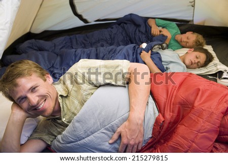 Family camping in tent, children (8-10) sleeping in background, focus on father resting in sleeping bag in foreground, smiling, portrait