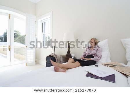Mature businesswoman sitting on bed with laptop, looking out window