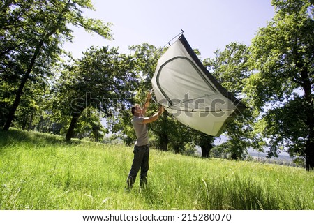 Young man lifting dome tent above head on camping trip in woodland clearing, side view (tilt)
