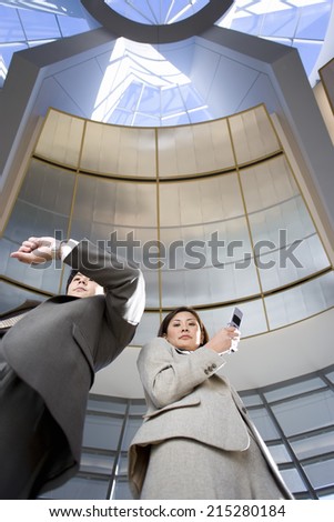 Businessman and woman standing below skylight in airport, man checking time on wristwatch, woman using mobile phone, portrait, low angle view