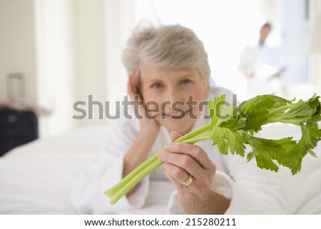 Senior woman lying on bed holding celery stick, smiling, portrait, close-up