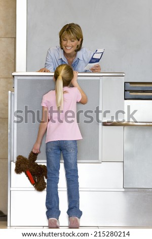 Girl , with soft toy, talking to female check-in attendant at airport check-in area, woman holding boarding passes, smiling, rear view